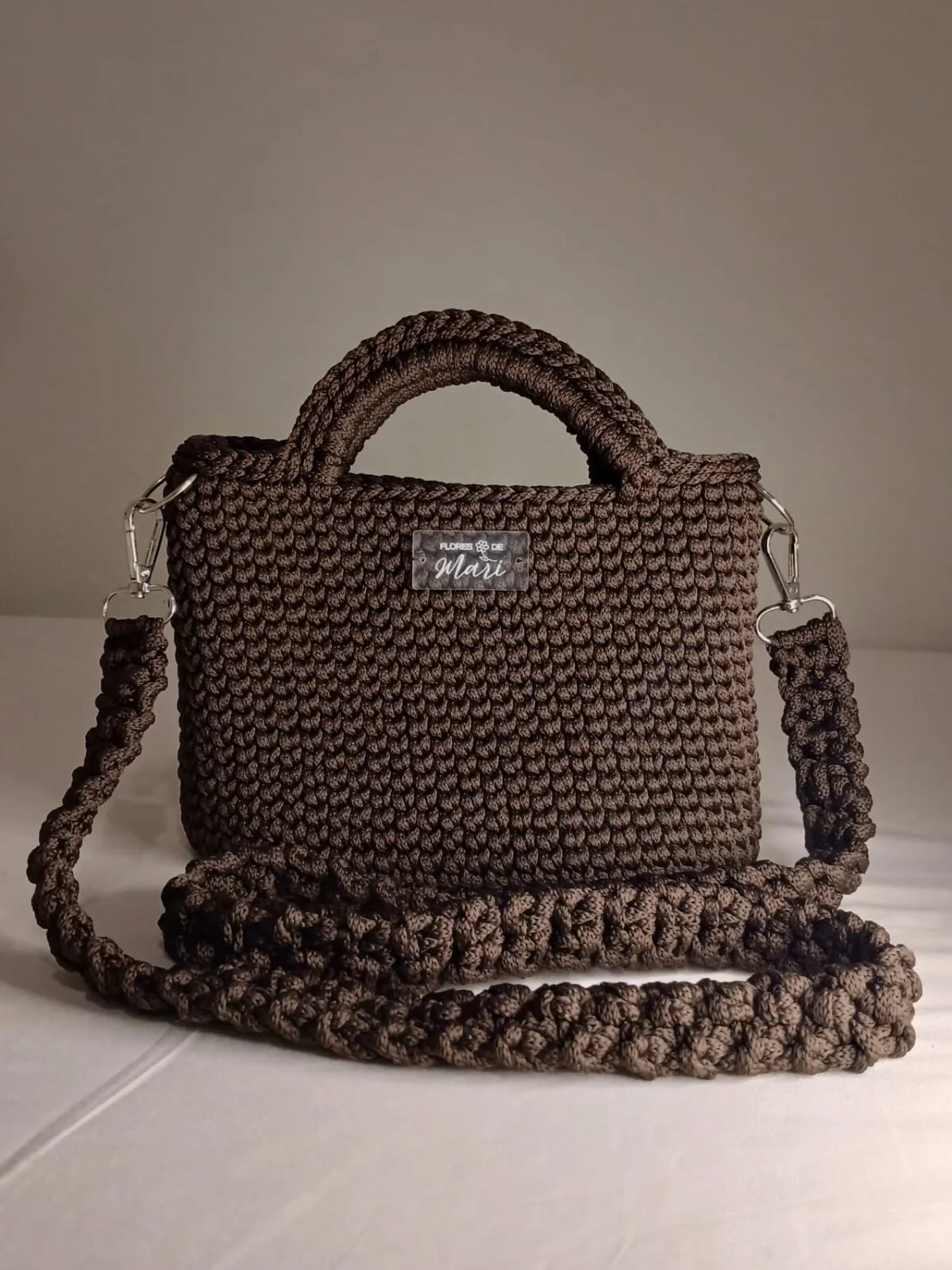 Bolsa de crochê marrom com duas alças de mão e alça transversal, etiqueta Flores de Mari na frente
