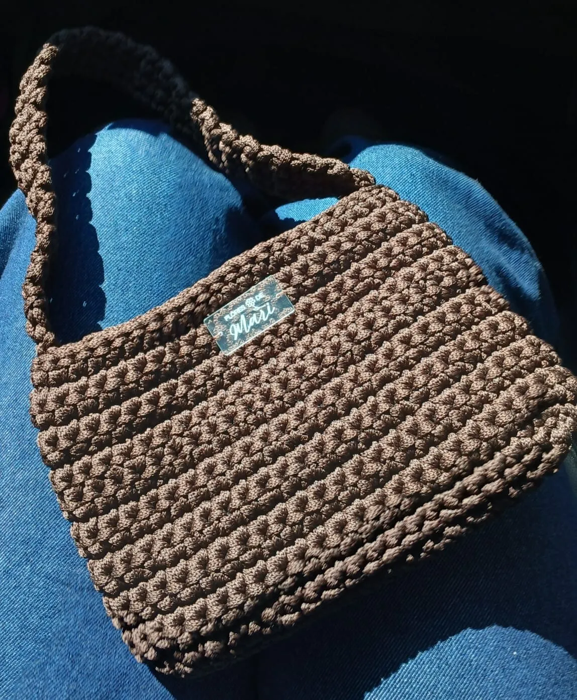 Bolsa de ombro de crochê marrom ao sol, com etiqueta Flores de Mari