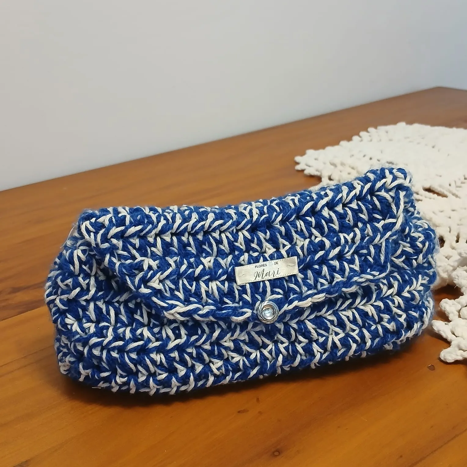 Clutch de crochê em azul e branco mesclado com botão prateado, sobre mesa de madeira