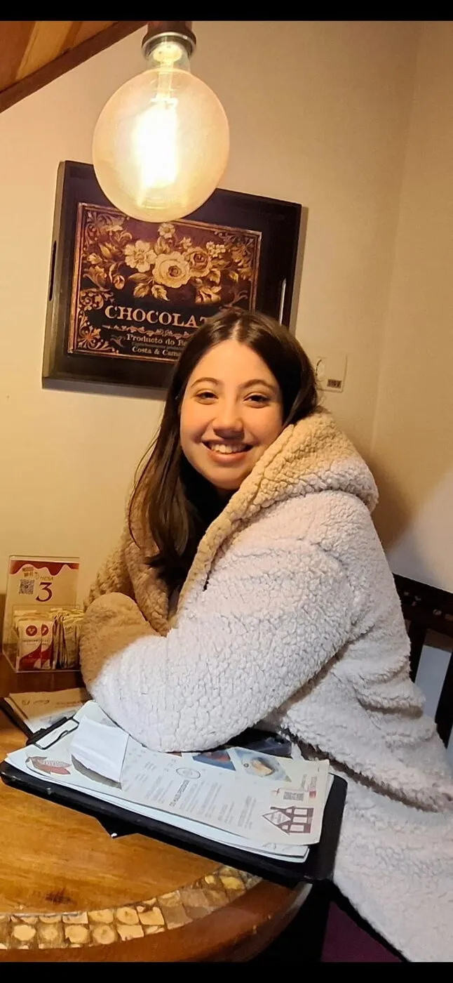 Mari sorrindo numa mesa de café, com um quadro de chocolate ao fundo