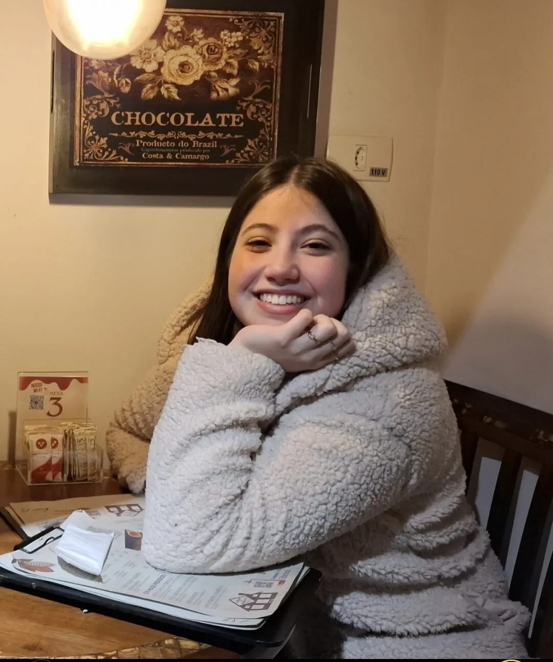 Mari sorrindo, apoiada numa mesa de café, com casaco de pelúcia claro