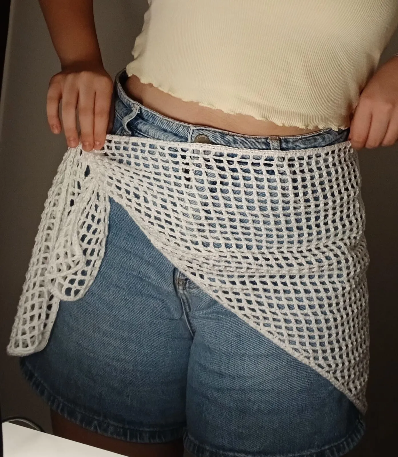 Saída de crochê branca em ponto rede, amarrada na cintura por cima de um short jeans