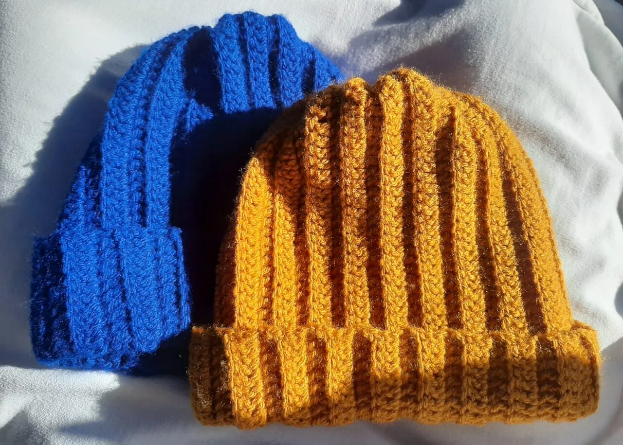 Duas toucas de crochê caneladas, uma azul royal e uma mostarda, sobre tecido branco